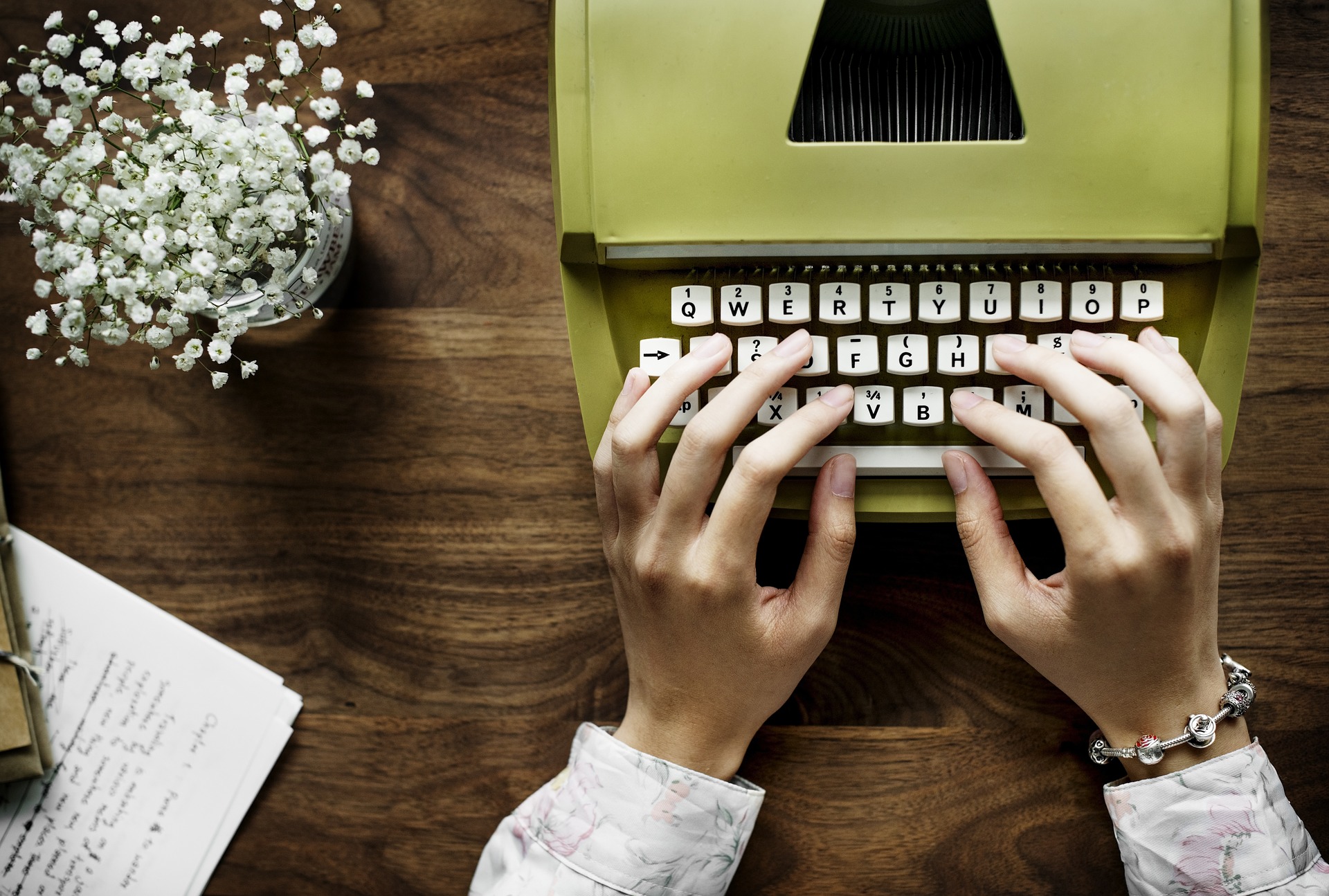 a person's hands poised at a typewriter and a container of flowers and written pages